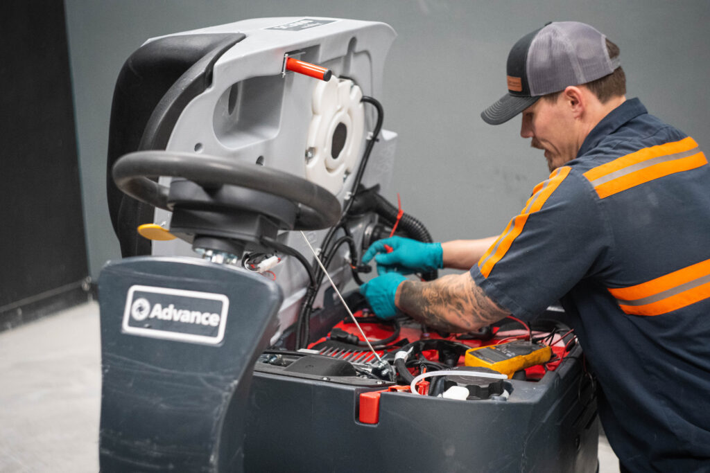 A WaySolutions technician performs electrical diagnostics on the open battery compartment of an Advance ride-on floor scrubber in Layton, Utah.

