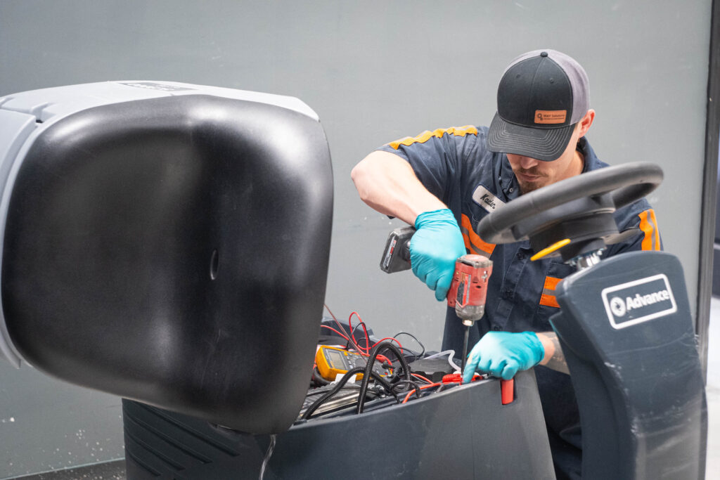 WaySolutions technician Kaden uses a cordless power drill to repair the internal battery compartment of an Advance ride-on floor scrubber.