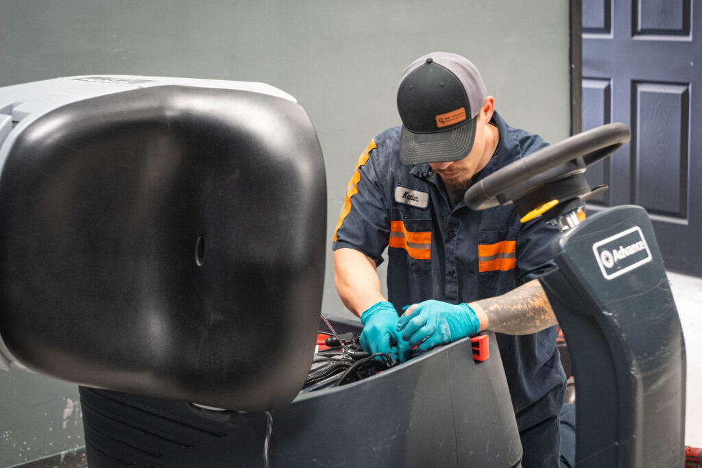 WaySolutions technician Kaden inspecting the internal electrical wiring and battery compartment of an Advance ride-on floor scrubber.
