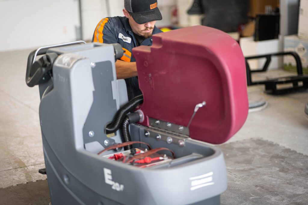A technician at WaySolutions inspects the open battery compartment of a Minuteman E20 commercial floor scrubber in Layton, Utah.