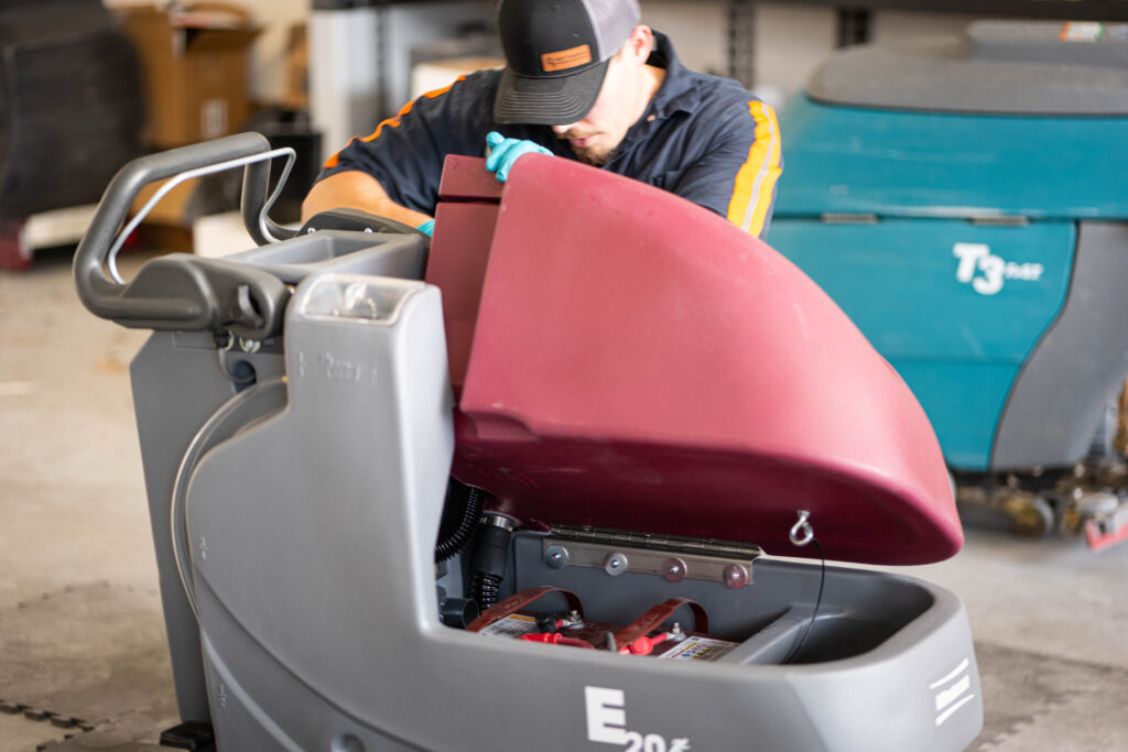 A WaySolutions technician inspects the internal battery compartment of a Minuteman E20 floor scrubber during a routine maintenance service.
