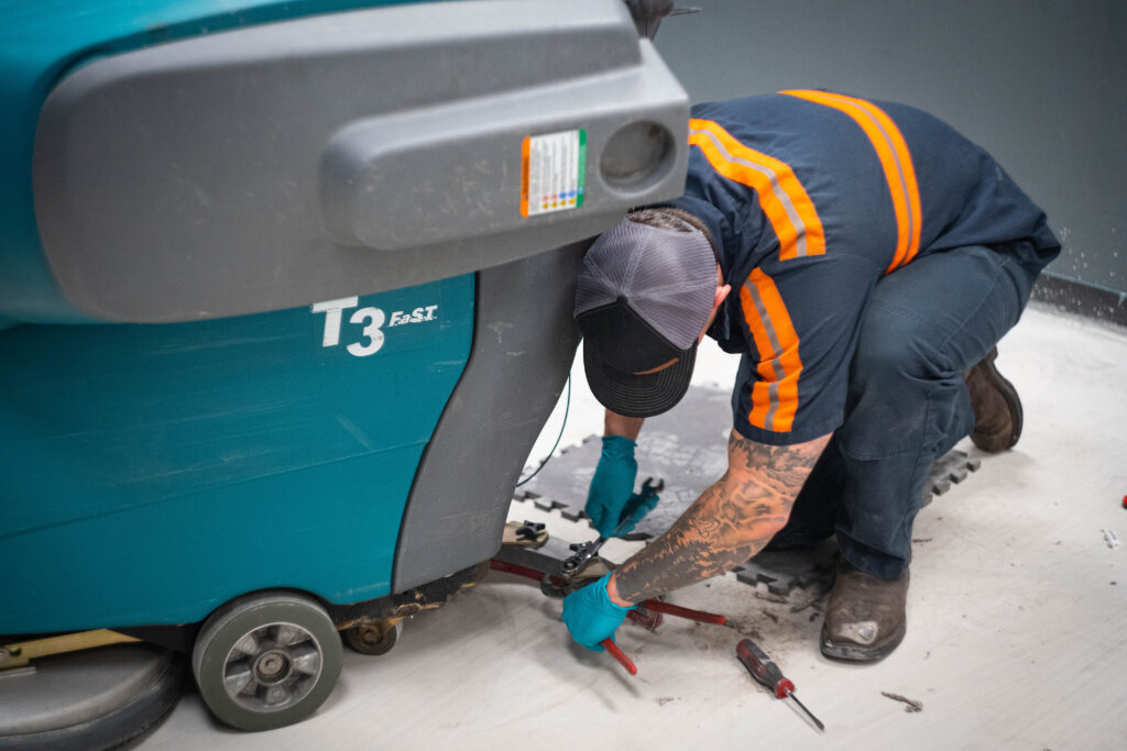A WaySolutions technician kneels to repair the undercarriage of a Tennant T3 walk-behind floor scrubber in Layton, Utah.
